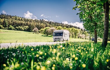 Weißes Wohnmobil fährt auf einer Strasse. Im Vordergrund ist eine Blumenwiese im Hintergrund eine Steuobstwiese mit angrenzendem Wald. | Odenwald | © Touristikgemeinschaft Odenwald e.V.