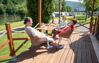 Es ist eine Holzterrasse zu sehen. Auf dieser sitzen 3 Frauen an einem Campingtisch. Dahinter stehen Wohnmobile auf einer Wiese