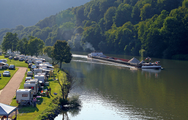 Ein Fluss (Neckar) mit einem Binnenschiff. Am rechten Ufer ist ein Campingplatz mit weißen Wohnwägen. Neckargemünd im Odenwald.
