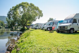 Auf der linken Bildseite fliesst ein Fluss. Auf dem Ufer rechts ist eine Wiese mit mehreren Wohmobilen. Im rechten hinteren Teil steht ein Baum.