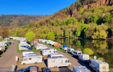 Campingplatz mit Wohnwägen und Wohnmobilen. Auf der rechten Seite ist ein Fluss (Neckar), dahinter am Ufer ein herbstlich bunter Wald. Neckargemünd im Odenwald