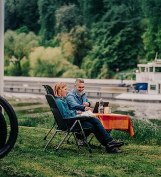 Ein Mann und eine Frau sitzen an einem Campingtisch vor Ihrem Wohnmobil am Ufer eines Flusses. Auf dem Fluß fährt ein Binnenschiff. Auf der anderen Uferseite ist Wald. Neckargemünd im Odenwald. | © Touristikgemeinschaft Odenwald e.V.