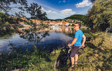 Ein Radfahrer mit blauem T-Shirt steht mit seinem Rad an einem Flussufer. Auf de anderen Uferseite sieht man eine Stadt. Eberbach im Odenwald. | © Touristikgemeinschaft Odenwald e.V.