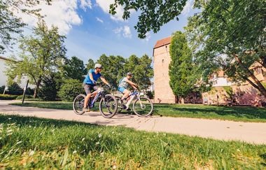 Zwei Radfahrer auf einem Radweg. Im Hintergrund ist eine Stadtmauer mit einem Turm. Eberbach im Odenwald. | © Tourstikgemeinschaft Odenwald e.V.
