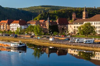 Im Vordergrund ist ein Fluß mit einem Binnenschiff. Dahinter am Ufer ist direkt ein Wohnmobilstellplatz mit 3 Wohnmobilen. Im Hintergrund ist eine Stadt mit Häusern und einer Kirche. Eberbach im Odenwald. | © Stadtverwaltung Eberbach