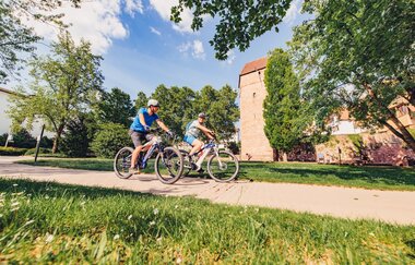 Zwei Radfahrer auf einem Radweg. Im Hintergrund ist eine Stadtmauer mit einem Turm. Eberbach im Odenwald. | © Tourstikgemeinschaft Odenwald e.V.