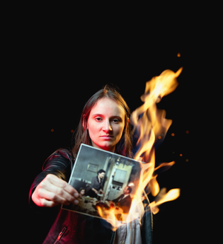 Eine junge Frau hält ein brennendes Foto in die Kamera. Auf dem Bild sieht man eine Familie am Küchentisch sitzen, die Mutter bedient den Vater.