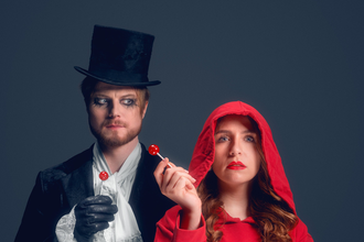 Links steht Herr Wolff mit einem schwarzen Mantel, einem schwarzen Zylinder und schwarzen Handschuhen, er trägt einen weißen Schal. Seine Augen sind schwarz umrandet. Er hat einen roten Lutscher in der rechten Hand und blickt verwirrt auf die Person neben