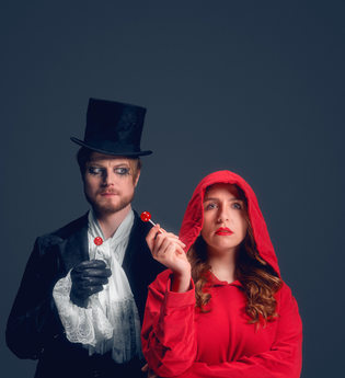 Links steht Herr Wolff mit einem schwarzen Mantel, einem schwarzen Zylinder und schwarzen Handschuhen, er trägt einen weißen Schal. Seine Augen sind schwarz umrandet. Er hat einen roten Lutscher in der rechten Hand und blickt verwirrt auf die Person neben