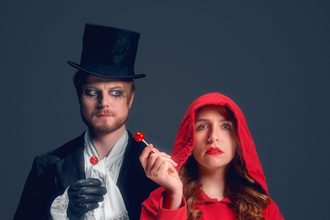 Links steht Herr Wolff mit einem schwarzen Mantel, einem schwarzen Zylinder und schwarzen Handschuhen, er trägt einen weißen Schal. Seine Augen sind schwarz umrandet. Er hat einen roten Lutscher in der rechten Hand und blickt verwirrt auf die Person neben