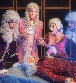 4 Personen mit Perücken stehen um einen Tisch auf dem eine weitere Person liegt