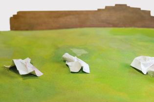 Drei zerknüllte weiße Zettel auf grünem Untergrund vor braunem Hintergrund.