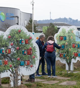 2 Besucher vor dem Gebäude der Klima Arena. Im Vordergrund zwei Plakate in BaumformPlakaten