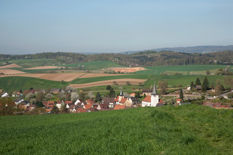 Blick auf das Dorf Spechbach mit mehreren Häusern und zwei Kirchen, umgeben von grünen Feldern und sanften Hügeln unter klarem Himmel. | © Landratsamt Rhein-Neckar-Kreis