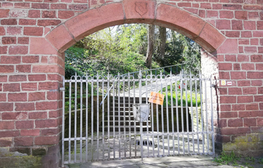 Ein Torbogen in einer roten Ziegelsteinmauer mit schmiedeeisernem Tor, ein Weg mit Treppen führt dahinter aufwärts in den Schlosspark. | © Stadt Weinheim - Jasmin Wolf