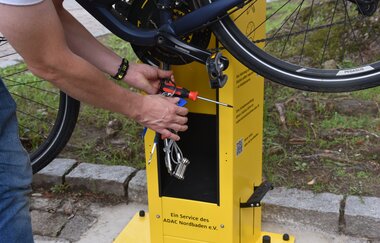 Gelbe Radservicestationauf der ein Fahrrad aufgehängt wurde und eine Person nach Fahrradwerkzeug greift. | © Bürgermeisteramt Oftersheim