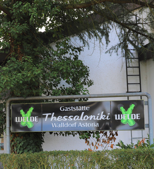 Schild "Gaststätte Thessaloniki" im Grünen vor einer weißen Wand