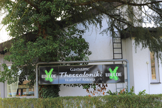 Schild "Gaststätte Thessaloniki" im Grünen vor einer weißen Wand