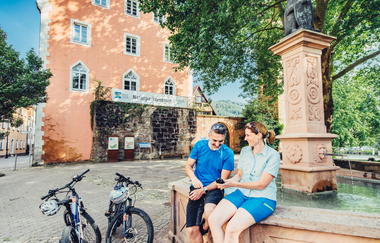2 Radfahrer sitzen an einem Brunnen und schauen in ein Handy. Vor dem Brunnen stehen die Fahrräder und ein blauer Rucksack. Im Hintergrund ist das Naturparkzentrum Eberbach zu sehen. | © Touristikgemeinschaft Odenwald e.V,
