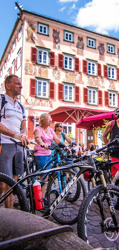 Mehrere Personen mit Fahrrädern stehen auf einem gepflasterten Platz vor einem historischen Fachwerkhaus mit roten Fensterläden und einem Café mit Sonnenschirmen. | © Landratsamt Rhein-Neckar-Kreis, Foto Adrian Weng