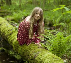 Kind mit langem Haar und gepunkteter Jacke sitzt auf moosbedecktem Baumstamm im Wald | © Landratsamt Rhein-Neckar-Kreis; Foto Dorothea Burkhardt