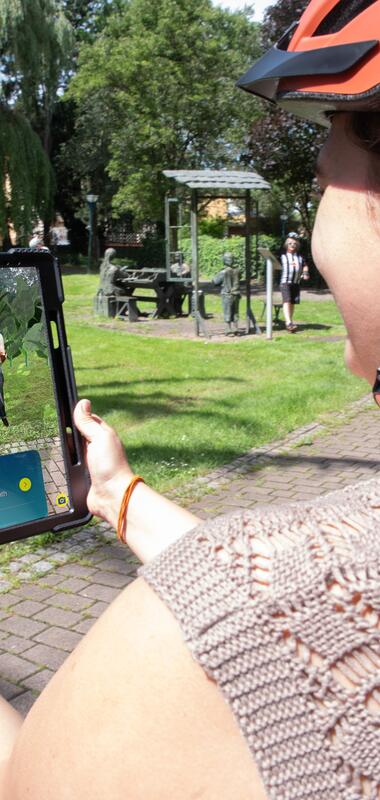 Frau mit orangefarbenem Helm hält Tablet, auf dem eine digitale Figur der Weitersehen App in einem Park angezeigt wird. | © Landratsamt Rhein-Neckar-Kreis; Foto Dorothea Burkhardt