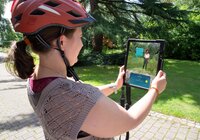 Frau mit orangefarbenem Helm hält Tablet, auf dem eine digitale Figur der Weitersehen App in einem Park angezeigt wird. | © Landratsamt Rhein-Neckar-Kreis; Foto Dorothea Burkhardt