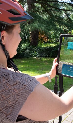Frau mit orangefarbenem Helm hält Tablet, auf dem eine digitale Figur der Weitersehen App in einem Park angezeigt wird. | © Landratsamt Rhein-Neckar-Kreis; Foto Dorothea Burkhardt
