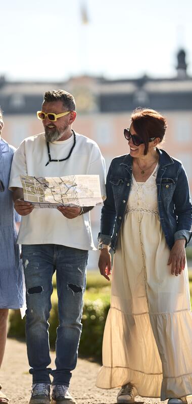 Drei Personen gehen auf einem Weg in einem Park vor dem Schwetzinger Schloss, eine Person hält eine Karte in der Hand. | © Rhein-Neckar-Kreis; Foto: Stefan Leitner